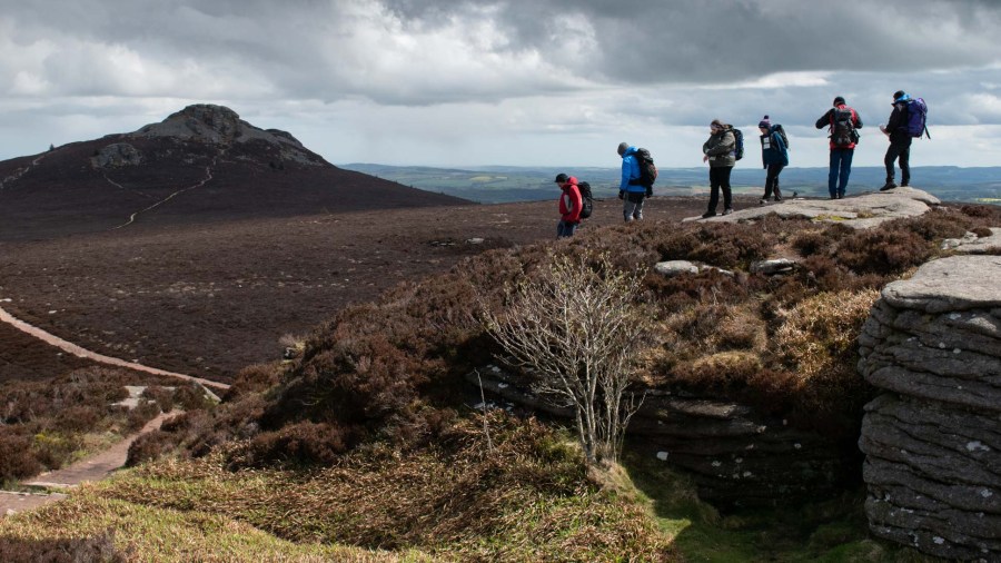 A group of adults on high moorland dressed in outdoor clothing being led towards the peak of Bennachie in the distance